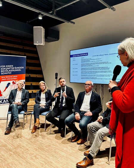 Fünf Personen in einer Podiumsdiskussion, eine Frau steht mit Mikrofon, Projektor zeigt Ablaufsplan.