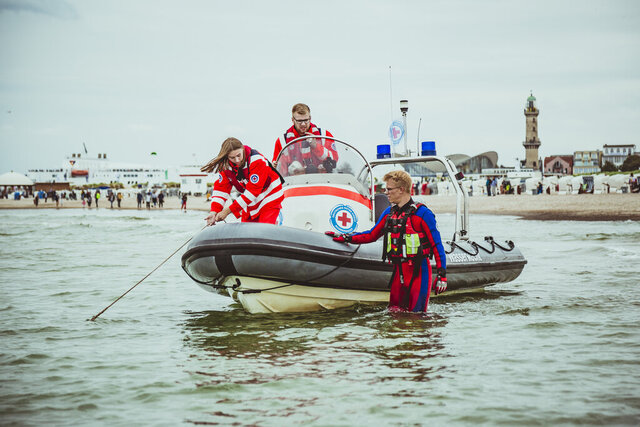 Zwei Rettungssanitäter in rot-weißen Uniformen auf einem Rettungsboot mit Rotkreuz-Logo, ein Kind in Neoprenanzug steht im Wasser daneben