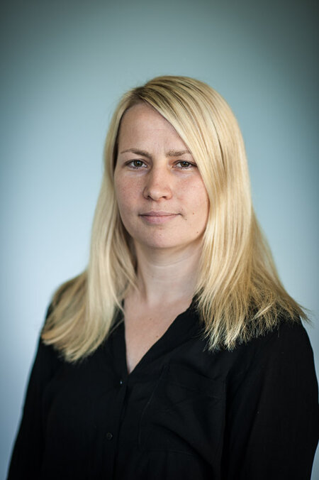 Frau mit langen blonden Haaren und schwarzem Oberteil vor neutralem Hintergrund.