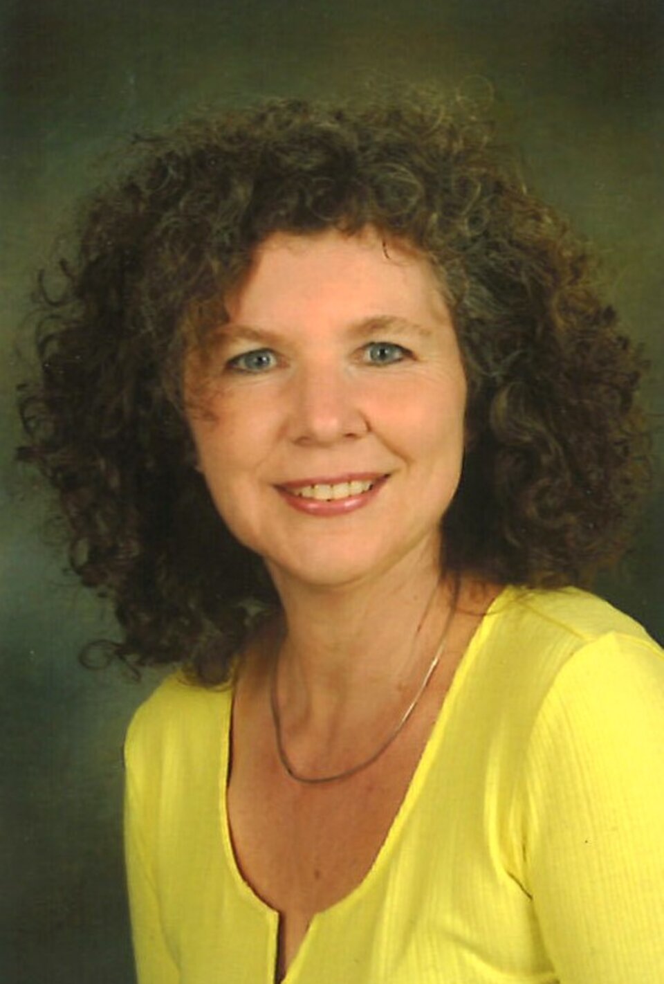 Frau mit lockigen Haaren und gelbem Oberteil lächelt vor einem verschwommenen, dunklen Hintergrund.