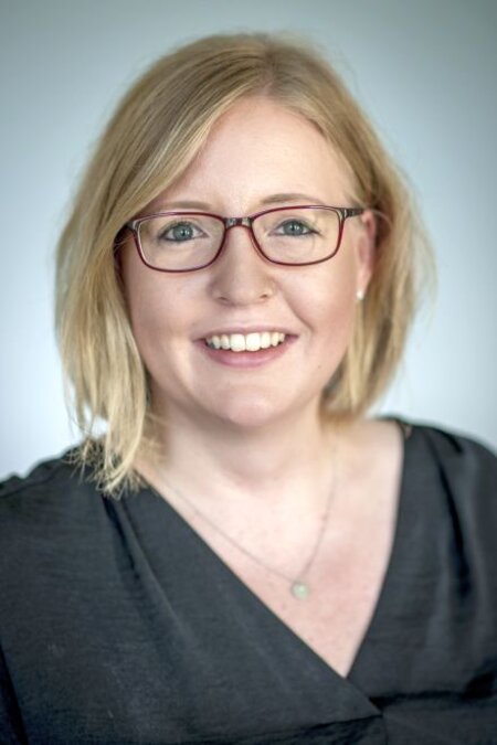 Person mit blonden Haaren und Brille lächelt in die Kamera. Sie trägt ein dunkles Oberteil.