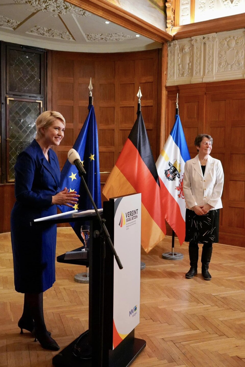 Zwei Frauen bei einer Rede, Flaggen von EU, Deutschland und Mecklenburg-Vorpommern im Hintergrund.