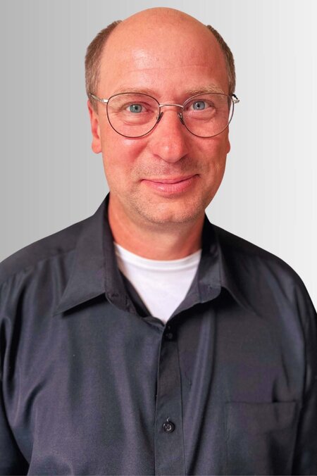 Mann mit Brille lächelt in die Kamera. Er trägt ein dunkles Hemd. Hintergrund ist hellgrau.