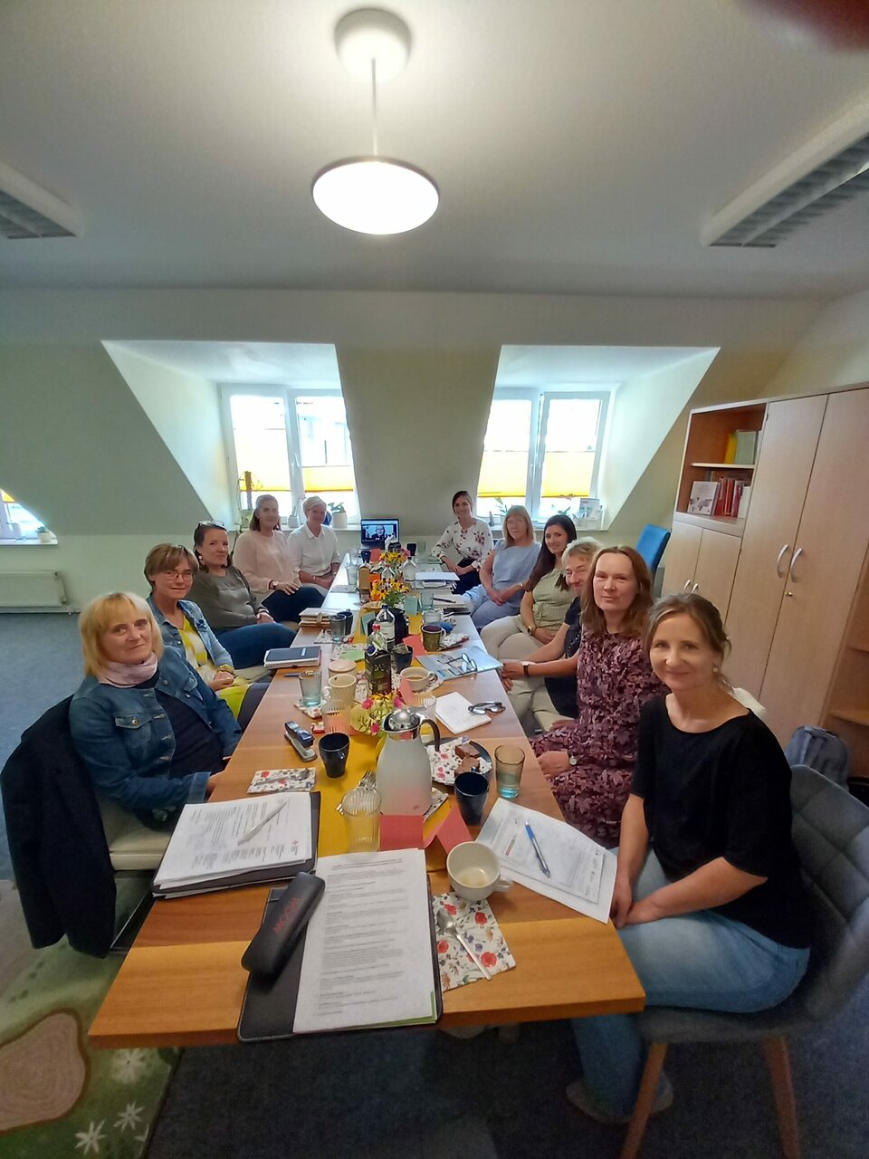 Elf Personen sitzen um einen Konferenztisch mit Getränken und Dokumenten in einem Büro unter einem Deckenlicht.