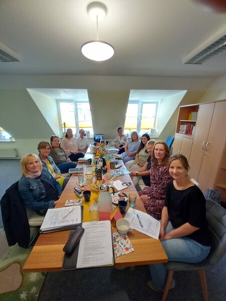 Elf Personen sitzen um einen Konferenztisch mit Getränken und Dokumenten in einem Büro unter einem Deckenlicht.