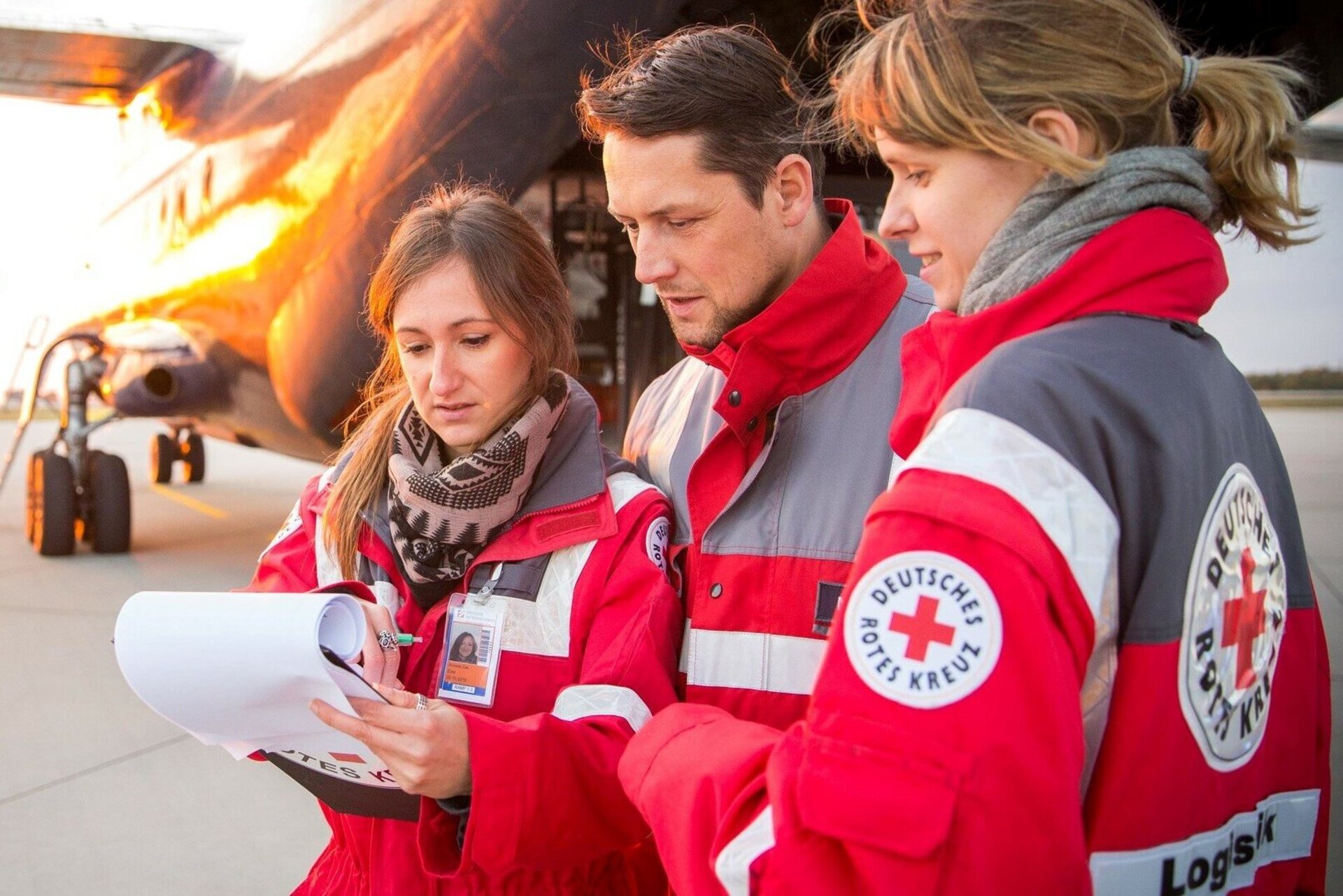 Drei Personen des Roten Kreuzes stehen vor einem Flugzeug und betrachten Dokumente bei Sonnenuntergang.