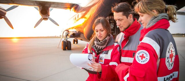 Drei Personen des Roten Kreuzes stehen vor einem Flugzeug und betrachten Dokumente bei Sonnenuntergang.