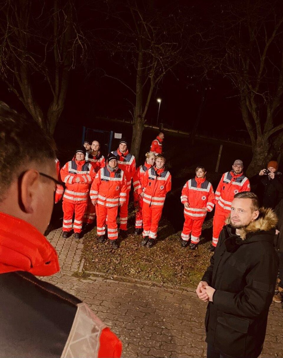 Gruppe von Menschen in roten Uniformen im Freien bei Nacht, zwei Personen vorne im Gespräch.