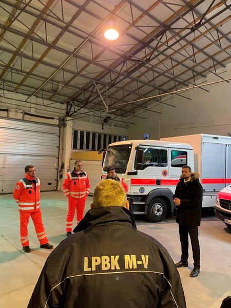 Vier Personen in Schutzkleidung und ein Mann in schwarzem Mantel vor Einsatzfahrzeugen in einer Halle.