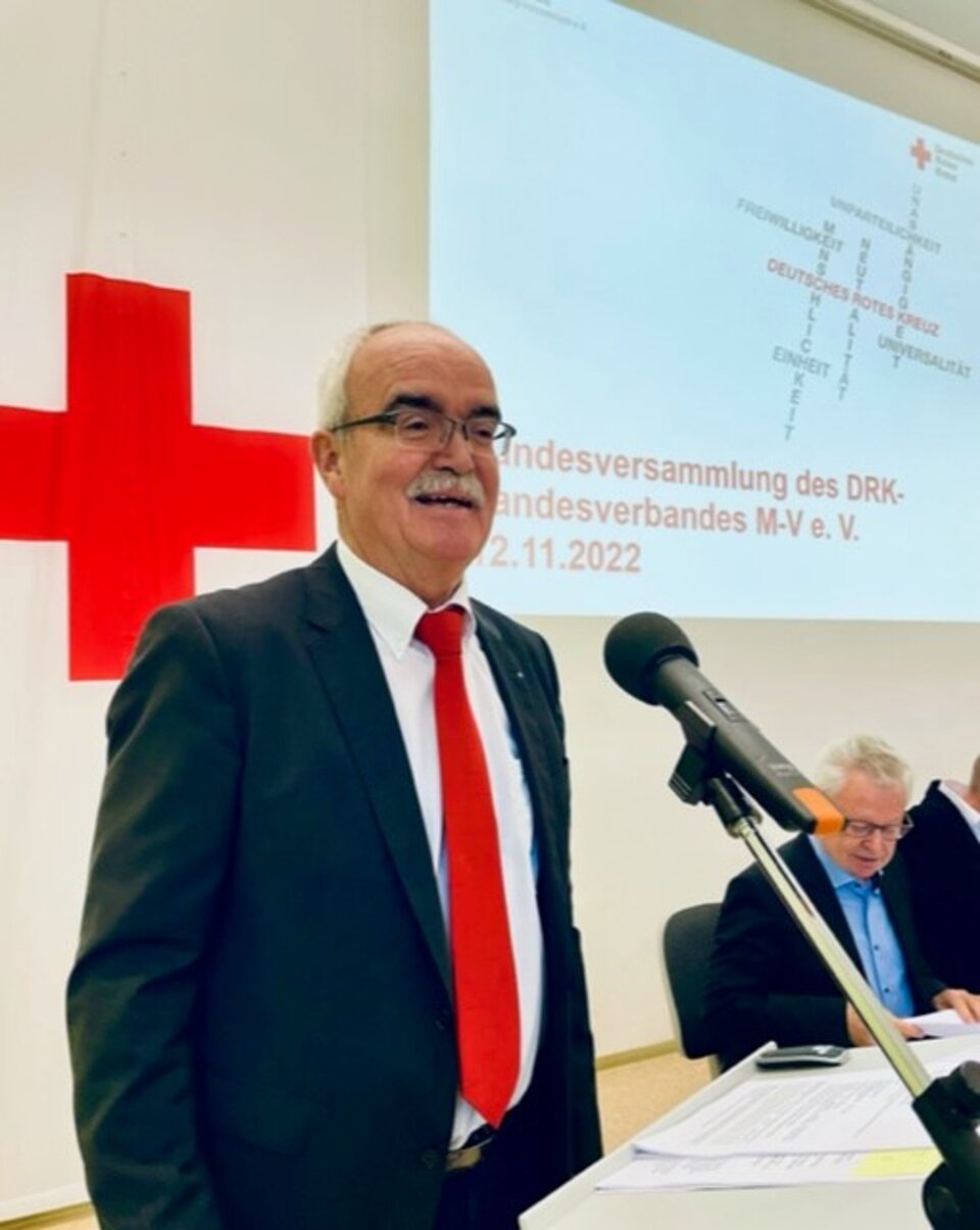 Mann in Anzug spricht bei DRK-Landesversammlung. Präsentation und rotes Kreuz im Hintergrund.