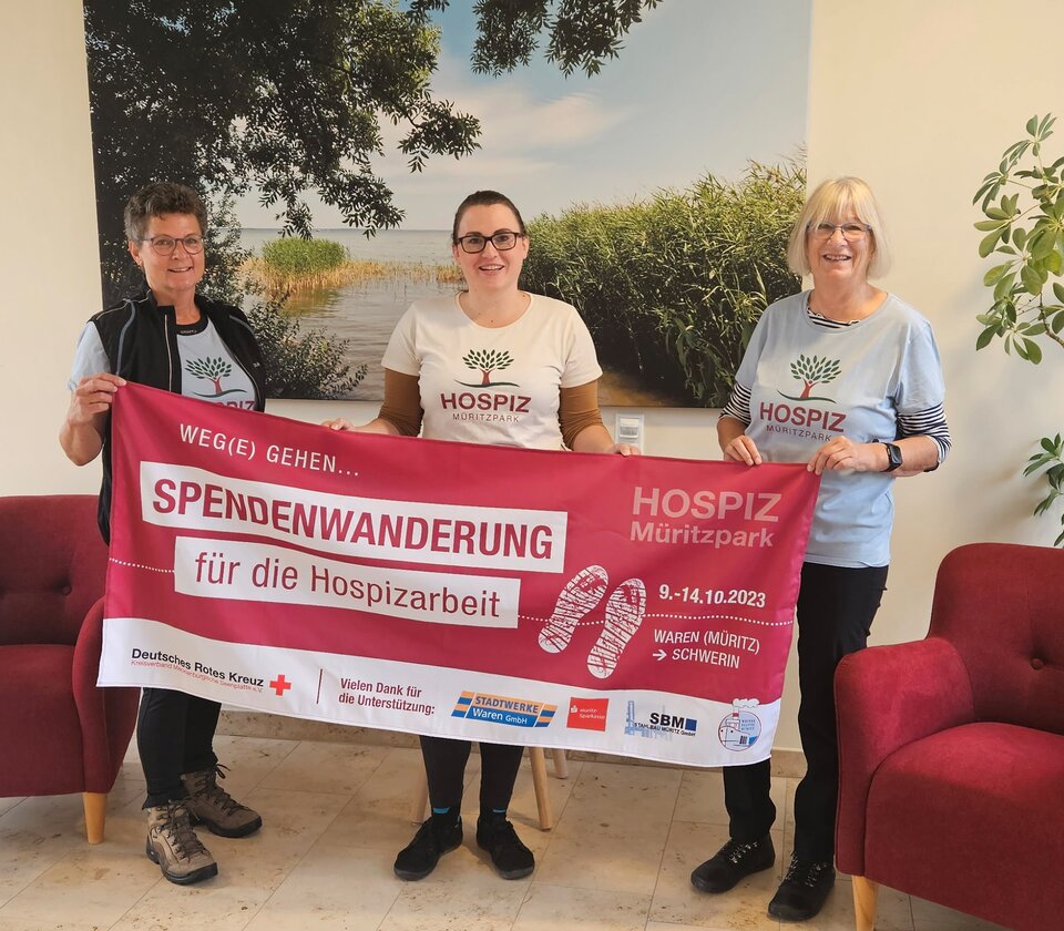 Drei Personen halten ein Banner zur Spendenwanderung für Hospizarbeit, im Hintergrund ein Landschaftsfoto.