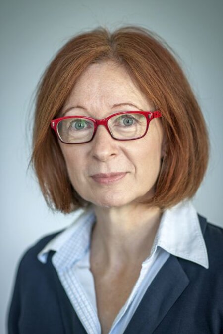 Frau mit roten Haaren und roter Brille, sieht in die Kamera, trägt Hemd und Blazer.