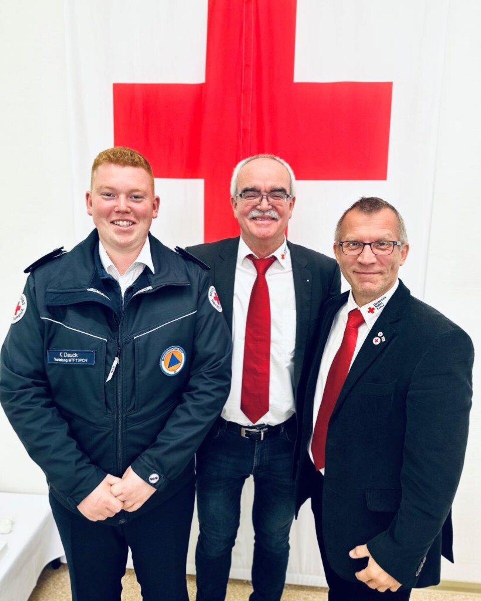 Drei Männer in Rotkreuzuniformen vor einem großen roten Kreuz auf weißem Hintergrund.