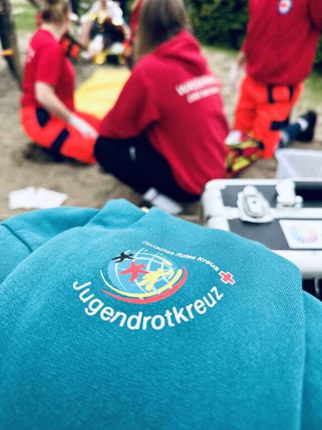 Logo des Jugendrotkreuz auf einem blauen Pullover, im Hintergrund Personen in Rotkreuz-Uniformen bei einer Übung