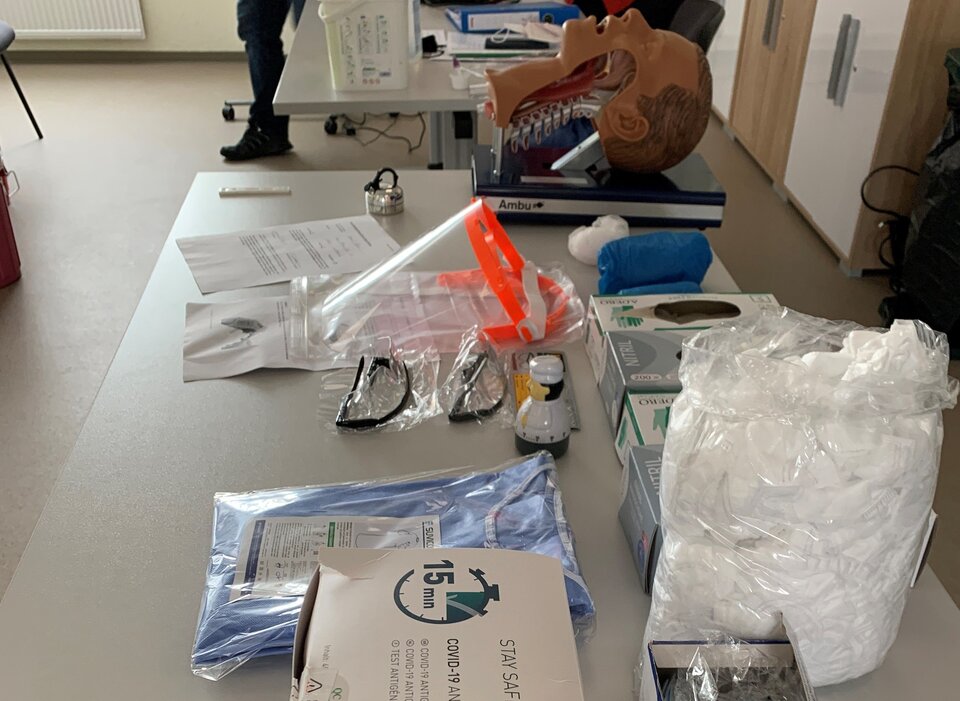 Tisch mit COVID-19-Testkits, Schutzbrille, Handschuhen und anatomischem Modellkopf im Schulungsraum.