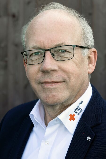 Mann mit Brille und Hemd mit "Deutsches Rotes Kreuz" Schriftzug, dunkler Anzug, neutraler Gesichtsausdruck.