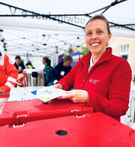 Lächelnde Frau in roter Jacke des Deutschen Roten Kreuzes an einem Verpflegungsstand unter einem weißen Pavillon.