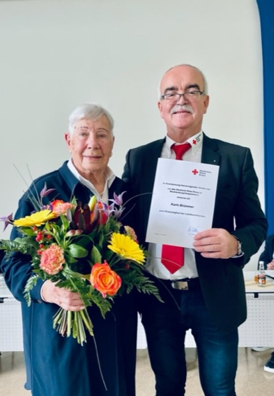 Ältere Frau mit Blumenstrauß und älterer Mann mit Urkunde. Beide lächeln in einem Raum vor einer Tafel.