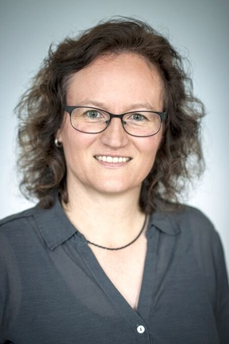 Person mit Brille und lockigem Haar lächelt freundlich in die Kamera, trägt ein dunkles Hemd.