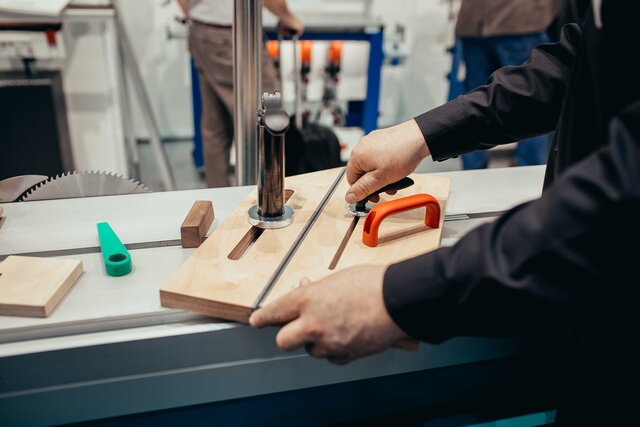 Person bedient Tischsäge mit Holzführung und rotem Griff in einer Werkstatt.