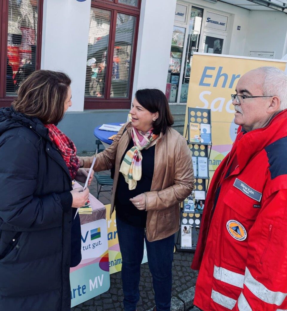 Drei Personen stehen vor einem Infostand mit Broschüren im Freien, einer in Rot mit Abzeichen, die anderen zwei in Jacken.