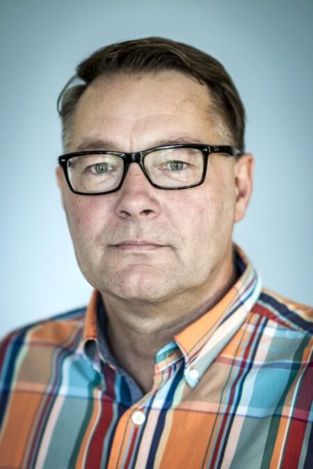 Mann mit Brille und bunt kariertem Hemd, ernst blickend, vor grauem Hintergrund.