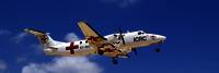 Weißes Flugzeug mit Rotkreuz-Logo in der Luft, blauer Himmel im Hintergrund.