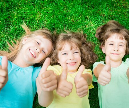 Drei lächelnde Kinder liegen auf Gras und zeigen Daumen hoch.