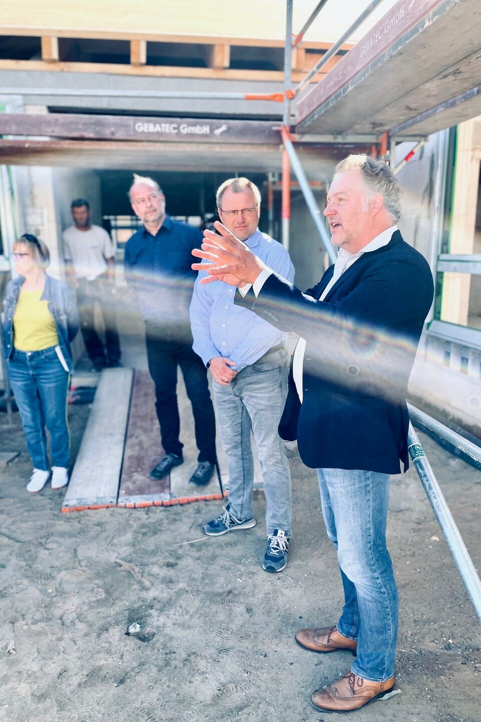 Mehrere Personen bei einer Baustellenbesichtigung, ein Mann spricht und gestikuliert, Baustellengerüst im Hintergrund.