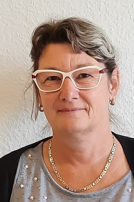 Person mit Brille vor einer hellen Wand, trägt eine Kette und ein graues Oberteil mit Perlendetails.