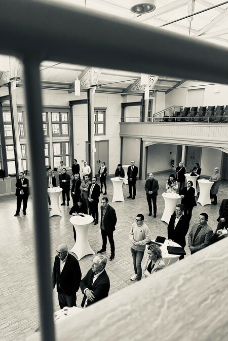 Personengruppe in einem großen Saal steht um Stehtische, Blick von oben, Geländer im Vordergrund. Schwarz-weiß-Foto.