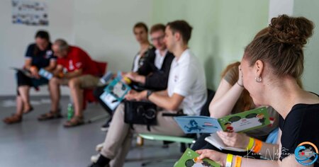 Personen sitzen in einem Raum in Reihe, lesen bunte Hefte und unterhalten sich.